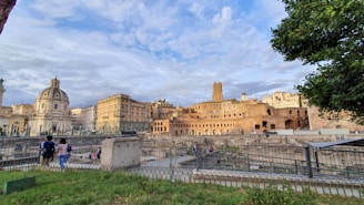 An expansive view of ancient Roman architecture with a mix of baroque and classical buildings featuring ornate details. In the foreground, a couple strolls near an excavation site surrounded by a metal fence. The sky is partly cloudy, and a tree frames the right side of the image.