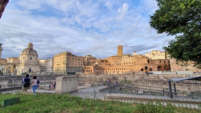 An expansive view of ancient Roman architecture with a mix of baroque and classical buildings featuring ornate details. In the foreground, a couple strolls near an excavation site surrounded by a metal fence. The sky is partly cloudy, and a tree frames the right side of the image.