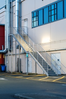 A metallic outdoor staircase ascends along the side of a light-colored industrial building with large blue-tinted windows. Several ventilation pipes run vertically along the building, and there are doors at the bottom of the staircase. The pavement in front of the building features yellow painted lines.