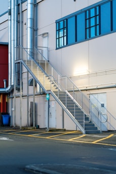 A metallic outdoor staircase ascends along the side of a light-colored industrial building with large blue-tinted windows. Several ventilation pipes run vertically along the building, and there are doors at the bottom of the staircase. The pavement in front of the building features yellow painted lines.