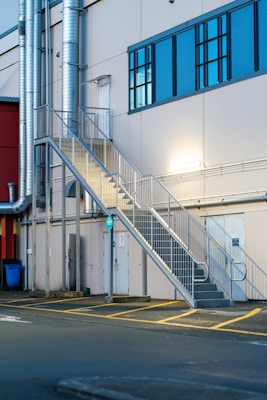 A metallic outdoor staircase ascends along the side of a light-colored industrial building with large blue-tinted windows. Several ventilation pipes run vertically along the building, and there are doors at the bottom of the staircase. The pavement in front of the building features yellow painted lines.