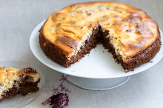 A homemade cheesecake with a swirled chocolate and cheese topping, presented on a white platter. A slice is missing, and a piece of the cake rests on a small glass plate nearby. There is a dried flower next to the glass plate, adding a rustic charm to the scene.