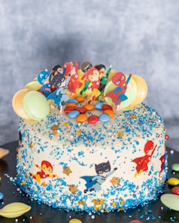 A decorated cake featuring superhero-themed edible figures and colorful candies. The cake is adorned with figures of various superheroes on top, surrounded by a sprinkle of blue and gold stars. Macarons in pastel colors are placed around the edges, complementing the vibrant color palette.