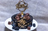 A richly decorated black cake is presented on a white plate. It is adorned with heart-shaped chocolates, gold foil accents, and small blackberries. A miniature whiskey bottle adds a unique touch to the top. A decorative "Happy Birthday" topper stands out against the cake's dark icing.