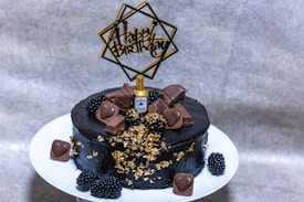 A richly decorated black cake is presented on a white plate. It is adorned with heart-shaped chocolates, gold foil accents, and small blackberries. A miniature whiskey bottle adds a unique touch to the top. A decorative "Happy Birthday" topper stands out against the cake's dark icing.