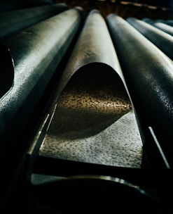 A detailed view of large metallic pipes with textured surfaces. The perspective is from below, highlighting the curvature and pattern of the pipes, with dramatic lighting creating shadows and contrasts.