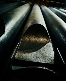 A detailed view of large metallic pipes with textured surfaces. The perspective is from below, highlighting the curvature and pattern of the pipes, with dramatic lighting creating shadows and contrasts.