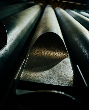 A detailed view of large metallic pipes with textured surfaces. The perspective is from below, highlighting the curvature and pattern of the pipes, with dramatic lighting creating shadows and contrasts.