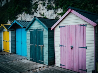 Four colorful beach huts are aligned side by side, each with vertical wooden planks and distinct colors: yellow, blue, teal, and pink. The huts have pitched roofs, and they stand on a wooden path with some pebbles. A white rock cliff with green vegetation is visible in the background.
