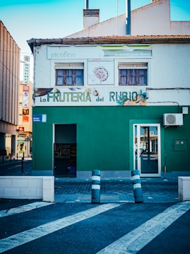 A small fruit store with a green facade, located on a street corner. Above the entrance, wooden window shutters are closed. The sign reads 'La Fruter&iacute;a del Rubio' with fruit images around it. A striped barrier pole stands at the entrance.