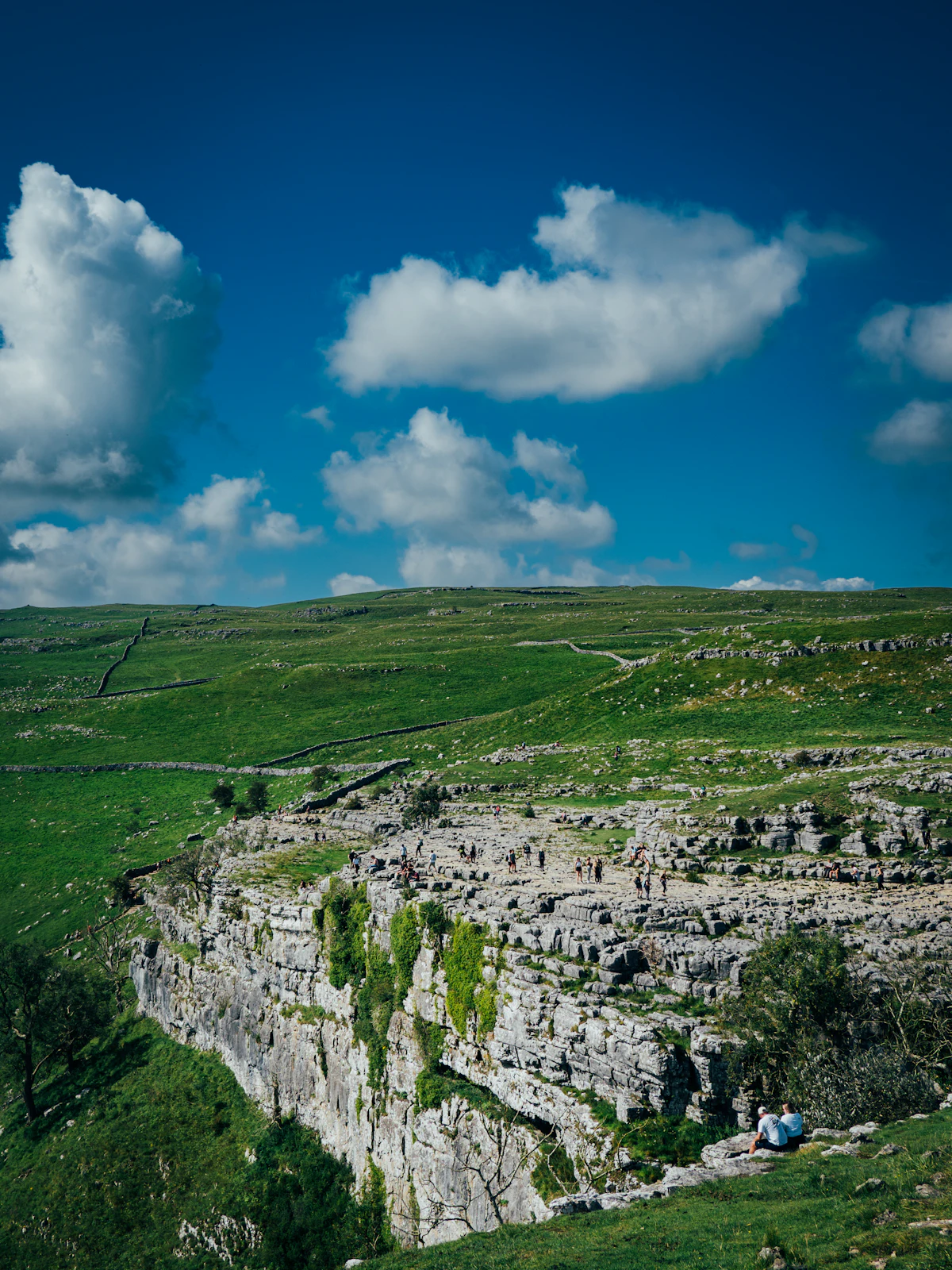 The curved limestone cliff of Malham Cove under a blue sky and white clouds