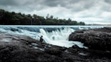 A serene landscape featuring a wide and gentle waterfall with misty water flowing over rocky edges. The scene is enveloped by dense green forest under an overcast sky. A lone individual sits calmly on the rocks near the waterfall, seemingly in contemplation.