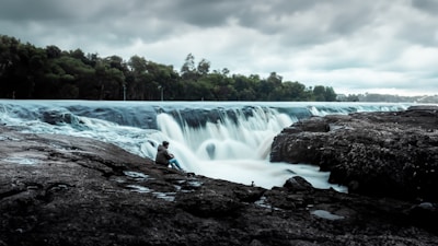 A serene landscape featuring a wide and gentle waterfall with misty water flowing over rocky edges. The scene is enveloped by dense green forest under an overcast sky. A lone individual sits calmly on the rocks near the waterfall, seemingly in contemplation.