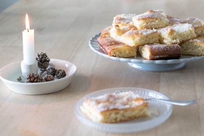 A plate of sugar-coated pastries rests on a wooden table. Nearby, a lit candle on a small dish is surrounded by pinecones, adding a cozy touch to the scene. A single piece of pastry is placed on a glass dish with a fork beside it.