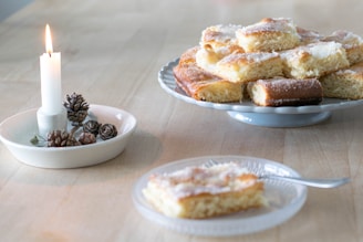 A plate of sugar-coated pastries rests on a wooden table. Nearby, a lit candle on a small dish is surrounded by pinecones, adding a cozy touch to the scene. A single piece of pastry is placed on a glass dish with a fork beside it.