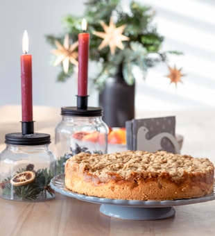 A cozy table setting with a homemade cake on a raised blue plate. Two red candles, lit and set in jar holders containing festive greenery and dried citrus slices, add warmth. A blurred background showcases a vase with evergreen branches and star decorations.
