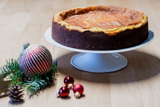 A cheesecake with a golden top and rich brown crust sits elegantly on a white cake stand. Nearby, festive decorations including a silver and red striped Christmas ornament and small red baubles rest on a wooden surface alongside green pine branches and a pine cone.