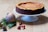 A cheesecake with a golden top and rich brown crust sits elegantly on a white cake stand. Nearby, festive decorations including a silver and red striped Christmas ornament and small red baubles rest on a wooden surface alongside green pine branches and a pine cone.