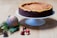 A cheesecake with a golden top and rich brown crust sits elegantly on a white cake stand. Nearby, festive decorations including a silver and red striped Christmas ornament and small red baubles rest on a wooden surface alongside green pine branches and a pine cone.