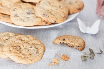 A plate of freshly baked chocolate chip cookies is set on a light-colored tablecloth. One cookie has a bite taken out of it, with crumbs scattered nearby. A small white decorative butterfly and some dry leaves are placed on the table for aesthetic effect.