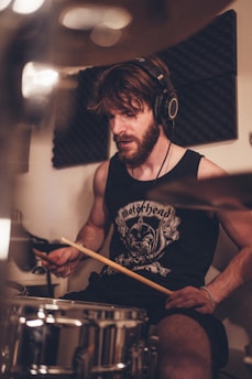 A bearded man is playing a drum set, wearing a sleeveless shirt with a band logo and headphones. The setting appears to be a soundproofed room, suggesting a music studio or rehearsal space. He is holding drumsticks, focused on his playing.