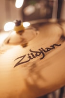 A close-up of a cymbal with the brand name 'Zildjian' clearly visible. The lighting creates soft reflections and highlights on the metallic surface, adding warmth and depth to the image.