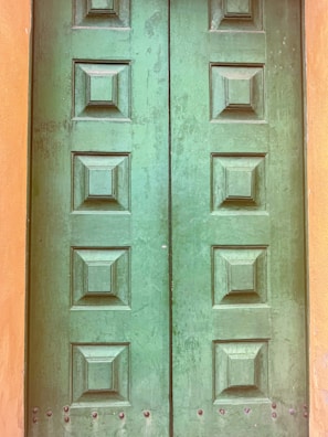 A pair of green wooden doors featuring a grid pattern with raised square panels. The wood appears slightly weathered, with some visible wear and tear, suggesting age. The frame around the door is painted a muted orange shade.