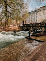 A group of people are gathered near a flowing river with rapids, surrounded by lush green trees and an elegant stone bridge. Some individuals are watching while others appear to be preparing to surf. In the background, a multi-story building with a classic architectural style stands prominently.