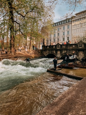 A group of people are gathered near a flowing river with rapids, surrounded by lush green trees and an elegant stone bridge. Some individuals are watching while others appear to be preparing to surf. In the background, a multi-story building with a classic architectural style stands prominently.