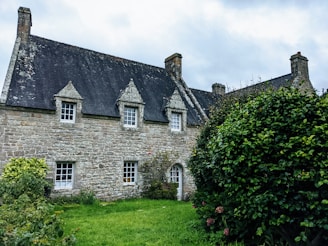 A rustic stone house with a steep slate roof and small attic windows. The property is surrounded by lush greenery and a well-maintained garden, with clouds visible in the overcast sky.