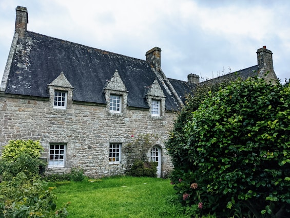A rustic stone house with a steep slate roof and small attic windows. The property is surrounded by lush greenery and a well-maintained garden, with clouds visible in the overcast sky.