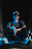 A person wearing a cap and glasses sits cross-legged on a surface, illuminated by blue lighting against a dark background. The individual wears a gray shirt and black pants with white sneakers, exuding a relaxed yet contemplative vibe.