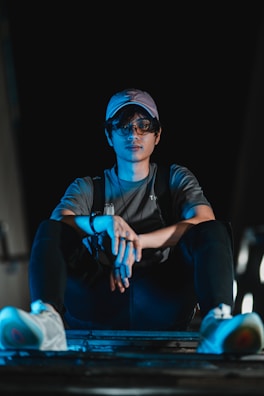 A person wearing a cap and glasses sits cross-legged on a surface, illuminated by blue lighting against a dark background. The individual wears a gray shirt and black pants with white sneakers, exuding a relaxed yet contemplative vibe.