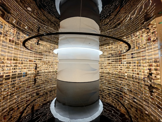 A futuristic library with a large, cylindrical pillar at its center. The walls are lined with shelves filled with books, and the design creates a mirrored illusion of endless space. The lighting is warm, adding a cozy atmosphere.