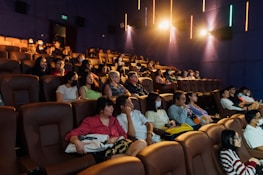 A group of people sit in a dimly lit theater, with some watching attentively while others wear masks. The seating consists of plush brown chairs and the atmosphere appears calm.