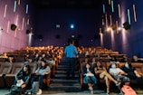 A group of people are seated in a theater with rows of comfortable brown seats and a man standing in front of them, possibly giving a presentation or lecture. The walls are purple with colorful vertical neon lights, and some attendees are wearing masks.