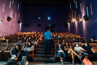 A group of people are seated in a theater with rows of comfortable brown seats and a man standing in front of them, possibly giving a presentation or lecture. The walls are purple with colorful vertical neon lights, and some attendees are wearing masks.