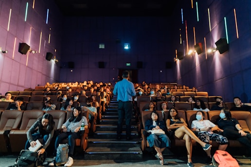 A group of people are seated in a theater with rows of comfortable brown seats and a man standing in front of them, possibly giving a presentation or lecture. The walls are purple with colorful vertical neon lights, and some attendees are wearing masks.