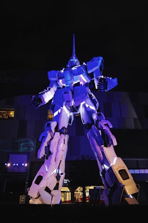 A large, illuminated robot statue stands prominently against a dark backdrop. The statue is highly detailed and constructed with angular panels and glowing elements, most notably lit in shades of blue and white. The architectural structure in the background adds a sense of scale and grandeur to the scene.