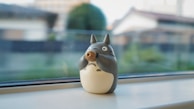 A small Totoro figurine is placed on a windowsill with a blurred background of greenery and a building outside. The figurine is gray and white with wide eyes and a pointed nose.