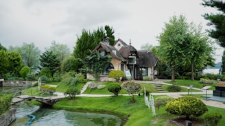 A quaint cottage surrounded by lush greenery, including various trees and shrubs. A stone pathway leads to the entrance, and a small bridge crosses a narrow stream in the foreground. The atmosphere is tranquil and inviting, resembling a scenic countryside landscape.