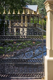 A decorative black wrought iron gate with intricate patterns stands in the foreground. Behind it, a pathway leads to a detailed, classic architectural structure with a veranda and ornate balustrades. Lush green foliage surrounds the area, providing a serene and inviting atmosphere.