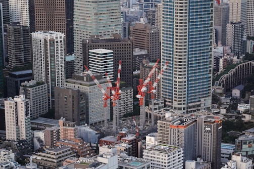 A dense urban landscape featuring numerous tall skyscrapers and a cluster of construction cranes in the midst of ongoing development. The buildings exhibit a variety of architectural styles, with glass, concrete, and metal facades. The cranes are striking with their red and white color, indicating active construction sites. The cityscape extends into the distance, showcasing a busy metropolitan area.