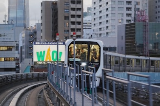 A modern train travels along an elevated track amidst an urban cityscape filled with tall buildings. In the foreground, the track curves slightly, and the train's front window reveals passengers inside. A large billboard displays the word 'WAO!' in bold letters.