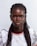 A person with braided hair is looking directly at the camera. They are wearing a white lacy top with colorful trim around the neckline. The person has earrings and a necklace with a pendant shaped like the African continent.