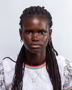 A person with braided hair is looking directly at the camera. They are wearing a white lacy top with colorful trim around the neckline. The person has earrings and a necklace with a pendant shaped like the African continent.