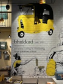 A mural on a tiled wall features a large yellow and black auto-rickshaw at the top. Below, there's a drawing of a woman in a traditional sari holding an ear of corn, surrounded by various vegetables. The word 'bhukad' is written with its Hindi script and definition — 'someone craving or consuming large quantities of food.' The setting includes a table and chairs beneath the artwork, suggesting a restaurant or café environment. There are decorative lights and framed pictures on the adjacent wall.