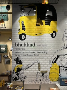 A mural on a tiled wall features a large yellow and black auto-rickshaw at the top. Below, there's a drawing of a woman in a traditional sari holding an ear of corn, surrounded by various vegetables. The word 'bhukad' is written with its Hindi script and definition — 'someone craving or consuming large quantities of food.' The setting includes a table and chairs beneath the artwork, suggesting a restaurant or café environment. There are decorative lights and framed pictures on the adjacent wall.