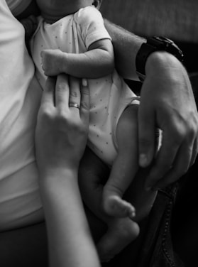 A close-up black and white image of a newborn baby being cradled, with a focus on the baby's small arm and legs. A hand with a ring gently rests on the baby's chest, conveying a sense of care and tenderness.