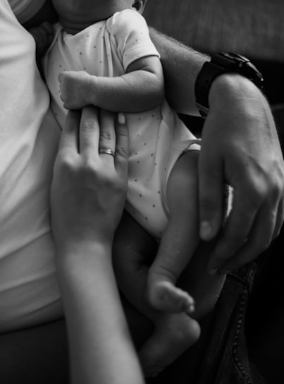 A close-up black and white image of a newborn baby being cradled, with a focus on the baby's small arm and legs. A hand with a ring gently rests on the baby's chest, conveying a sense of care and tenderness.